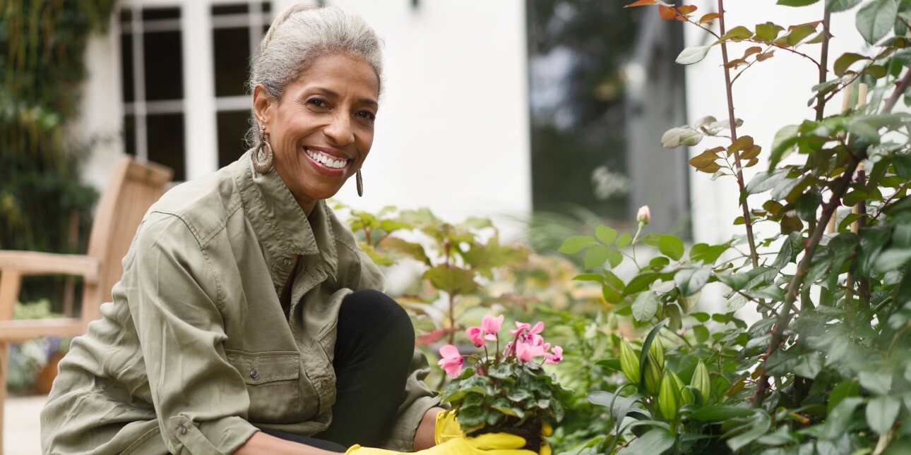 A smiling, older Black woman with grey hair, hooped earrings and khaki jacket, kneels down in her garden. Wearing yellow gloves, she is planting a pink flowering plant. Around her are green bushes a wooden bench and a house
