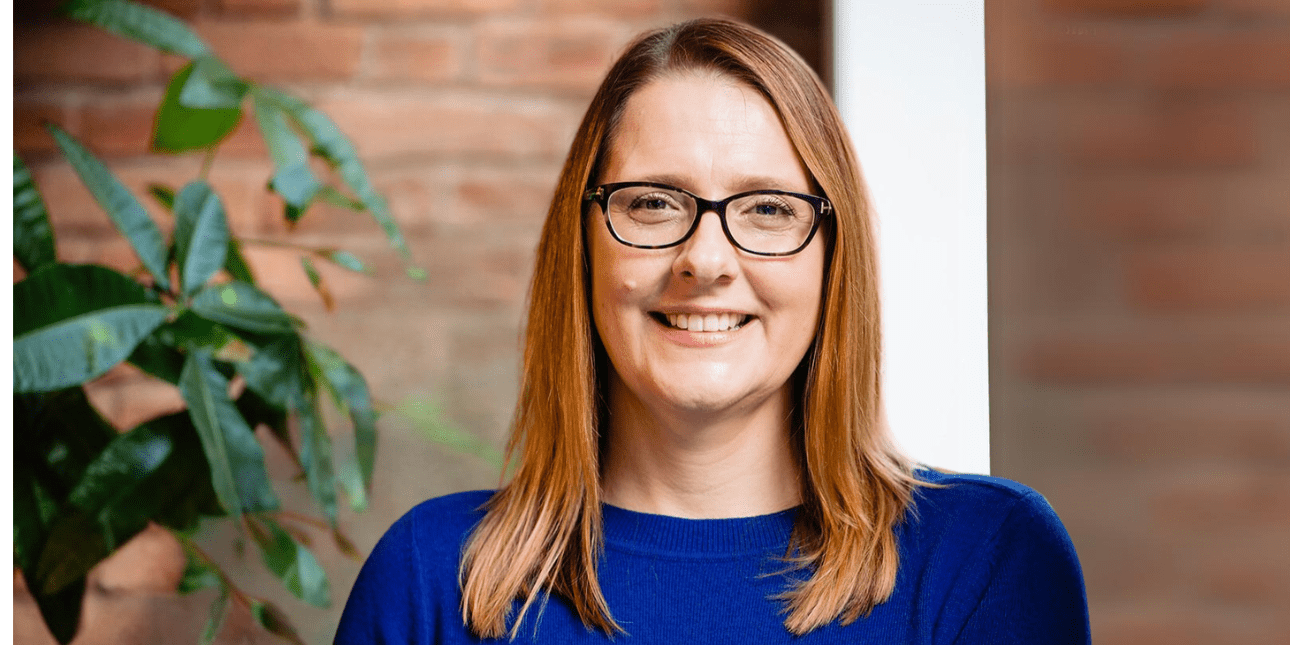 A portrait of a smiling Donna Castle, a white woman with shoulder length chestnut hair, wearing glasses and a blue jumper. There is a plant and brick wall in the background