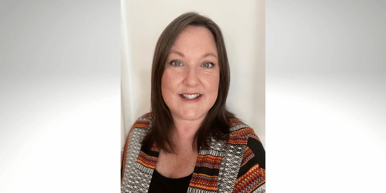 A portrait of Kelly Quigley-Hicks smiling at the camera. Kelly is a white woman with shoulder-length brown hair, wearing a brown and orange patterned jacket