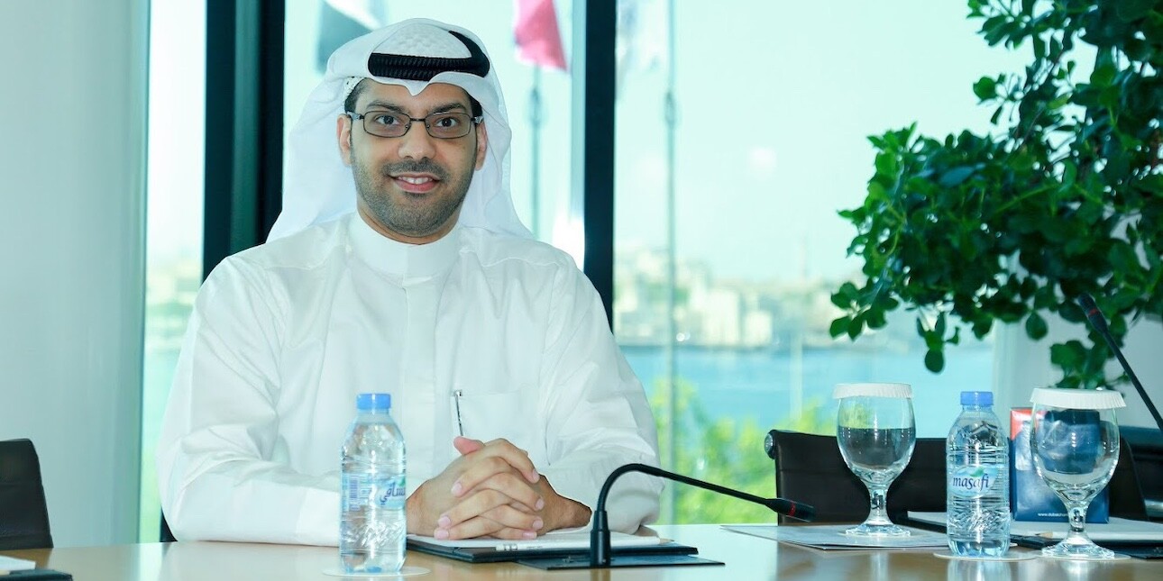Saad Al Rubaiaan wearing glasses and a white ghutra and thobe while sat at a desk with water bottles and glasses on it. He is in front of a window which has flags and greenery outside