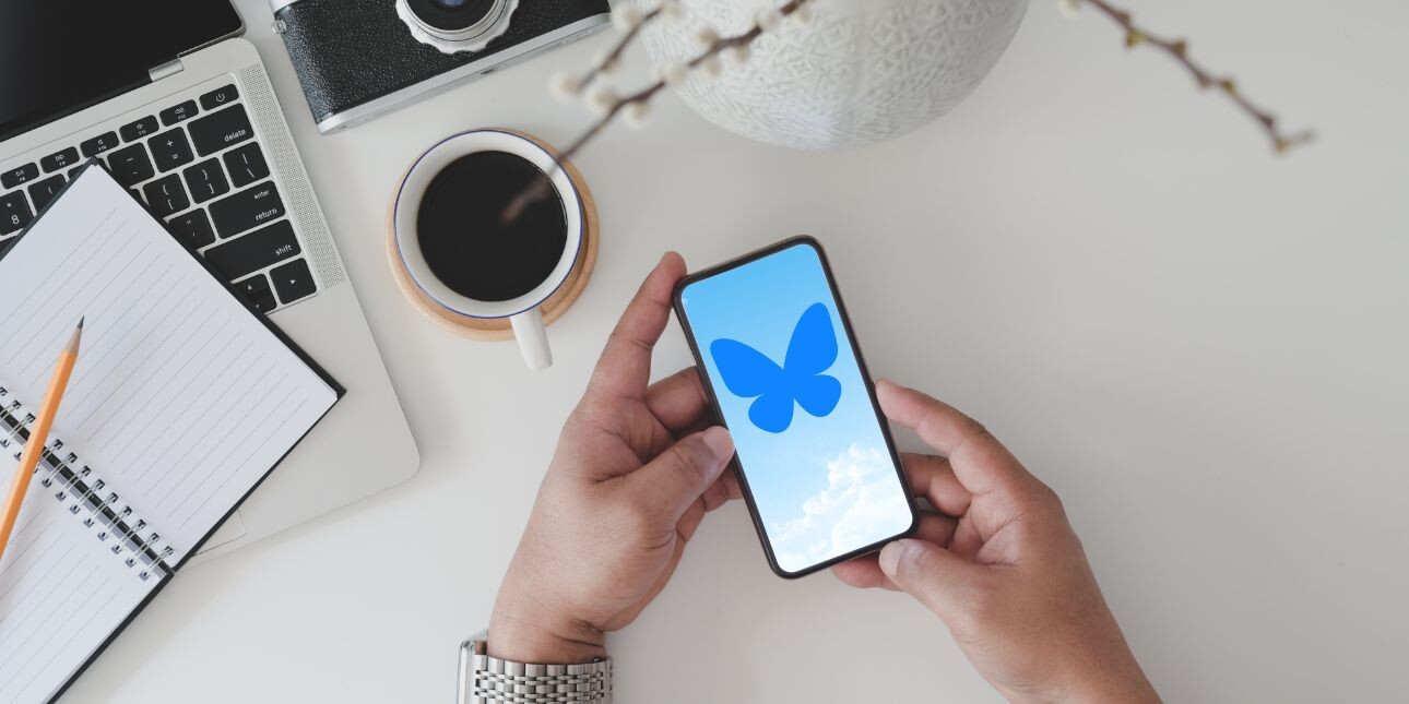 A pair of hands (with a watch on the left one) holds a mobile phone with an image of a blue butterfly on it and some clouds, in the background is a cup of coffee, a laptop, notepad and pencil.
