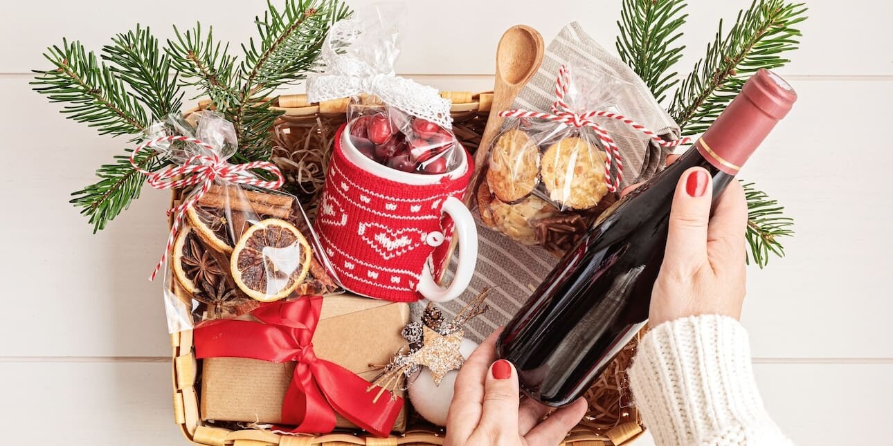 A Christmas hamper basket containing wrapped presents, ribbons, wooden spoons, biscuits and a mug covered in a red knitted cover. A white hand with red nail varnish lifts a bottle of red wine from the hamper