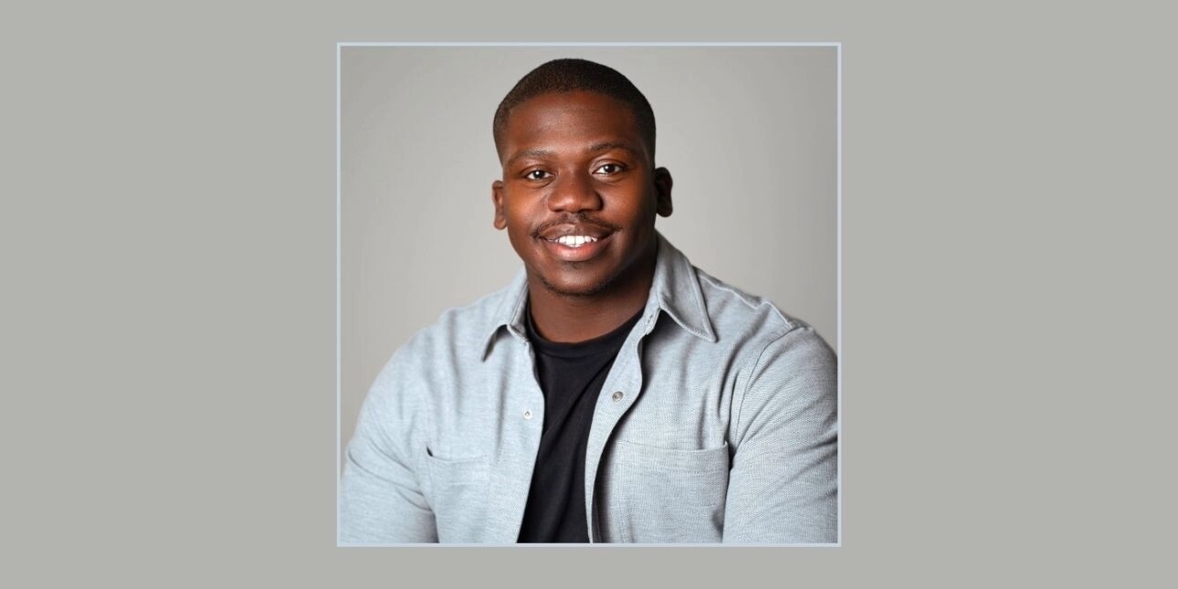 A portrait of Tariq Peters on a grey background. Tariq is a Black man with short hair and a light-grey shirt over a black t-shirt