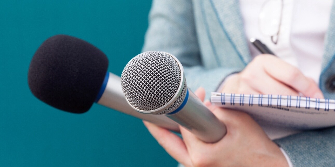 A close up of the torso and white hands of a journalist writing in a notebook while holding two microphones. Her suit is grey, the background is a turquoise