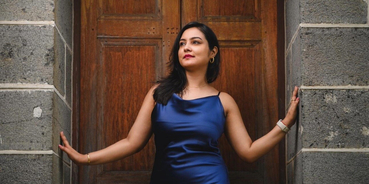 Hilla Lakhpatwala wearing a blue dress in front of wooden door with her arms reaching out to the stone porch. Hilla is an Asian woman with long dark hair.