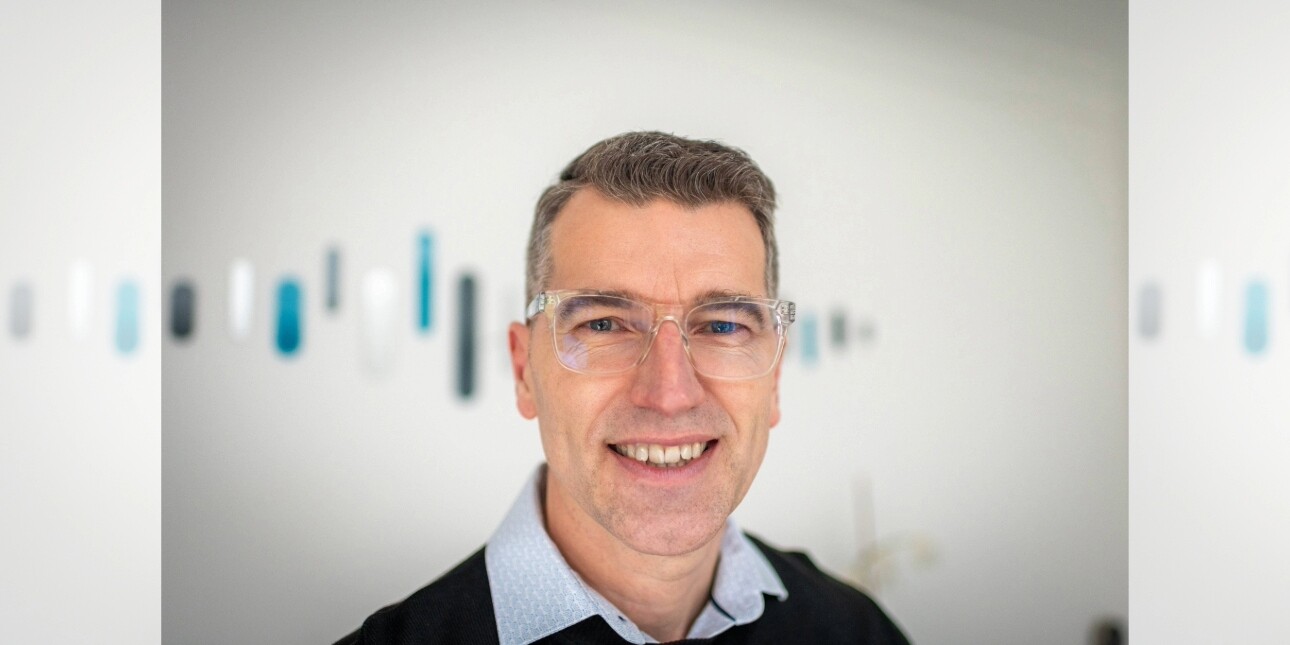 A portrait of Jon Large in front of a grey wall featuring blue-turquoise specks. Jon is a white man with short brown-grey hair, who wears transparent framed spectacles and blue collared shirt under a dark jumper