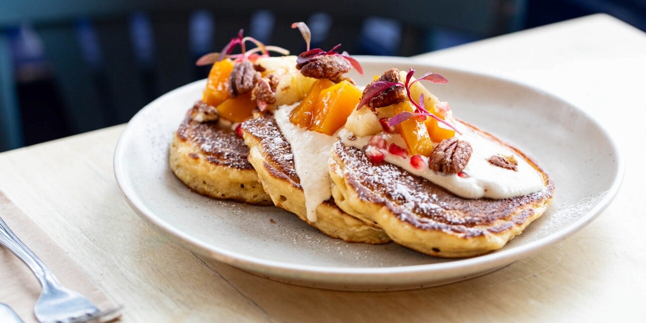 Three pancakes on a white plate on a table, piled with fruit and nuts