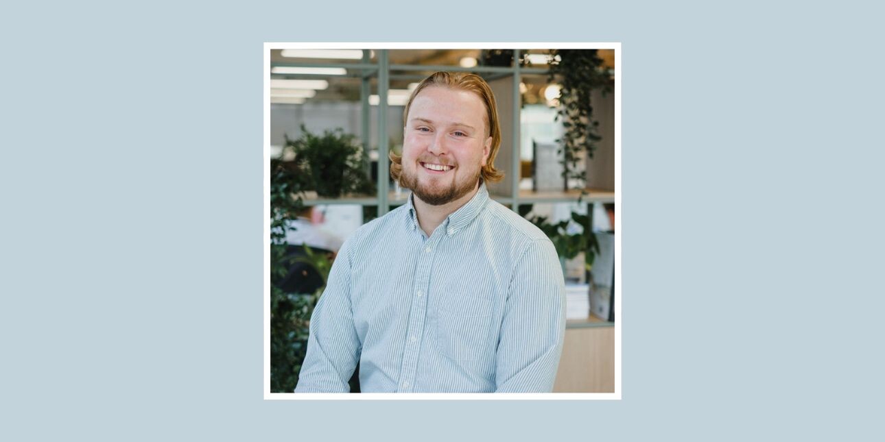 A white smiling man looks at the camera. He has ginger hair and a beard and is wearing a blue shirt and the background is an office