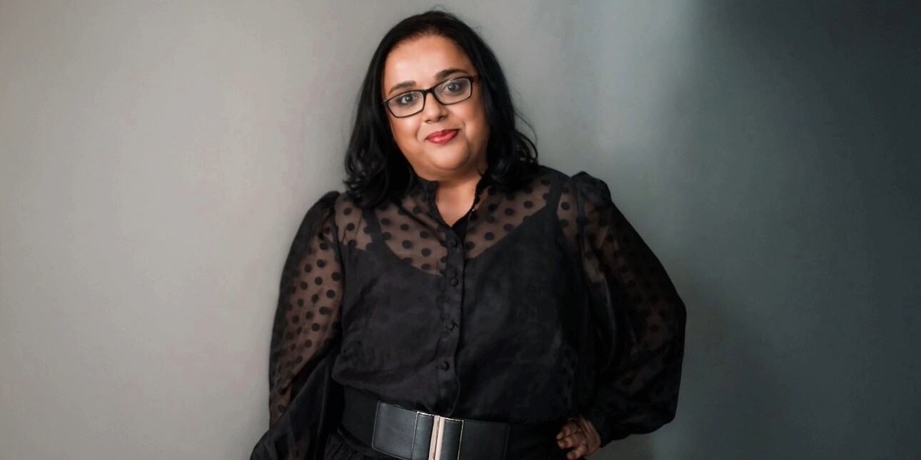 A colour portrait of Advita Patel against a grey backdrop. Advita is a British-Asian woman with shoulder length dark hair and glasses, who wears a black top.