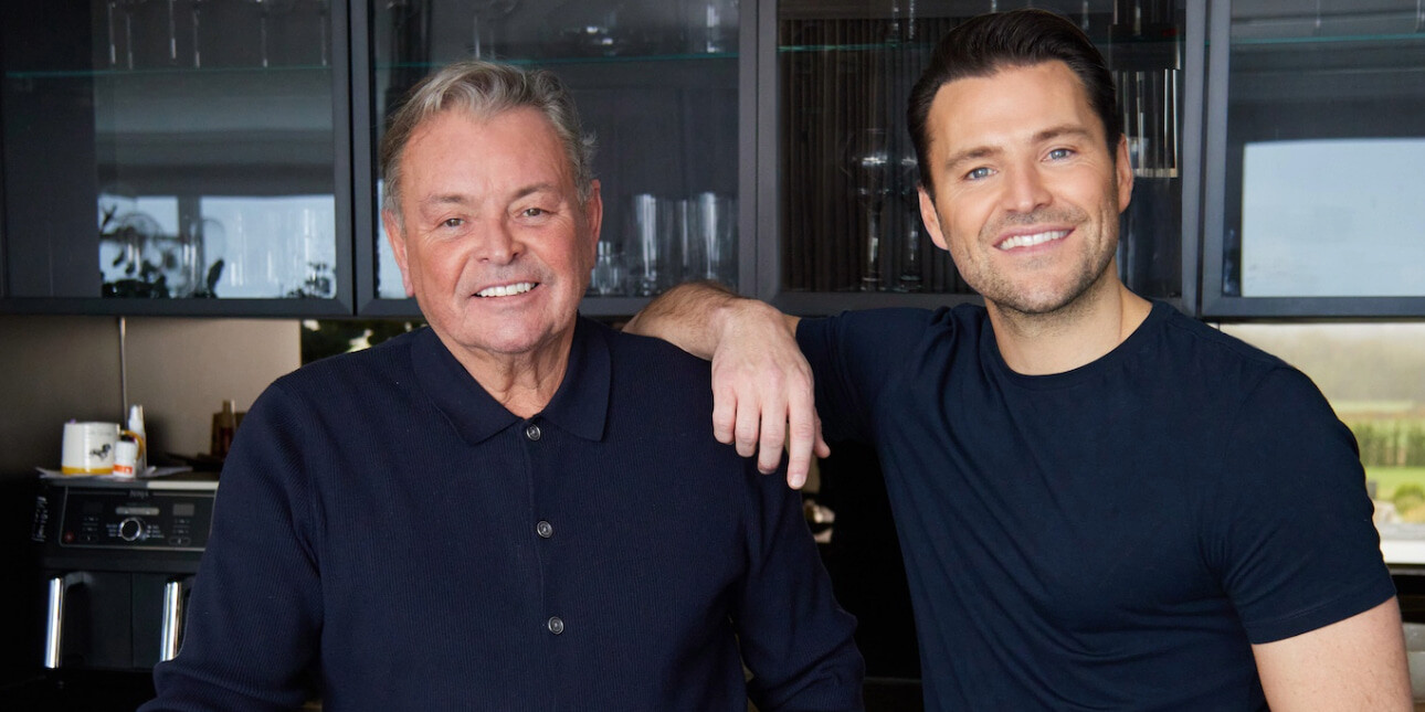 A colour photograph of Mark Wright Snr - a white man with grey hair, wearing a navy shirt - and his son Mark Wright - a white man with dark hair and navy t-shirt. They are stood in a dark kitchen, Wright Jr rest his right arm on his father's shoulder