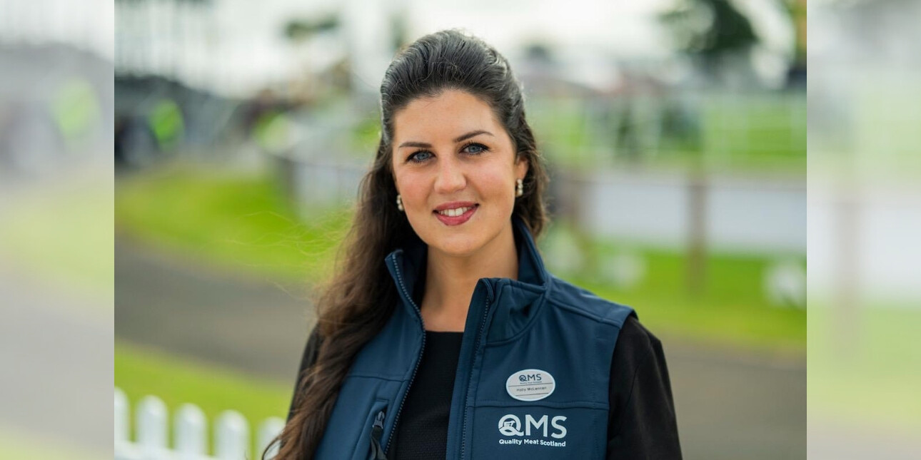 A colour portrait of Holly McLennan. Holly is a white woman with long dark hair wearing a blue gilet with writing on it. There is greenery behind her.