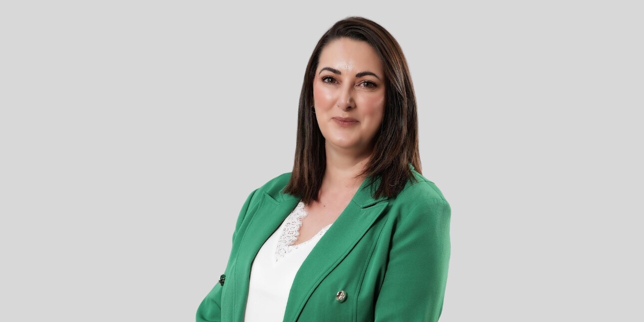 A colour portrait of Natasha Irvine against a grey background. Natasha is a white woman with shoulder length brown hair who wears a white top and green jacket. She smiles at the camera.