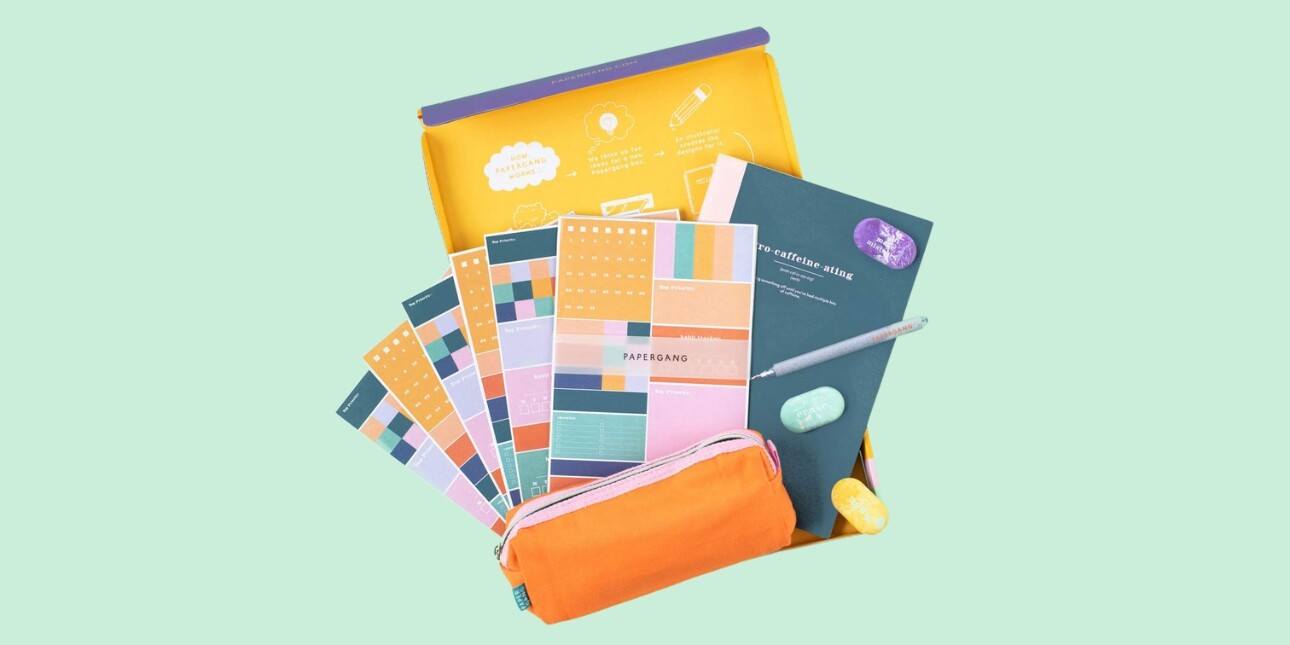 On a green pastel background, a birds eye view of an assortment of brightly coloured notebooks, planners, pencil case and other stationery