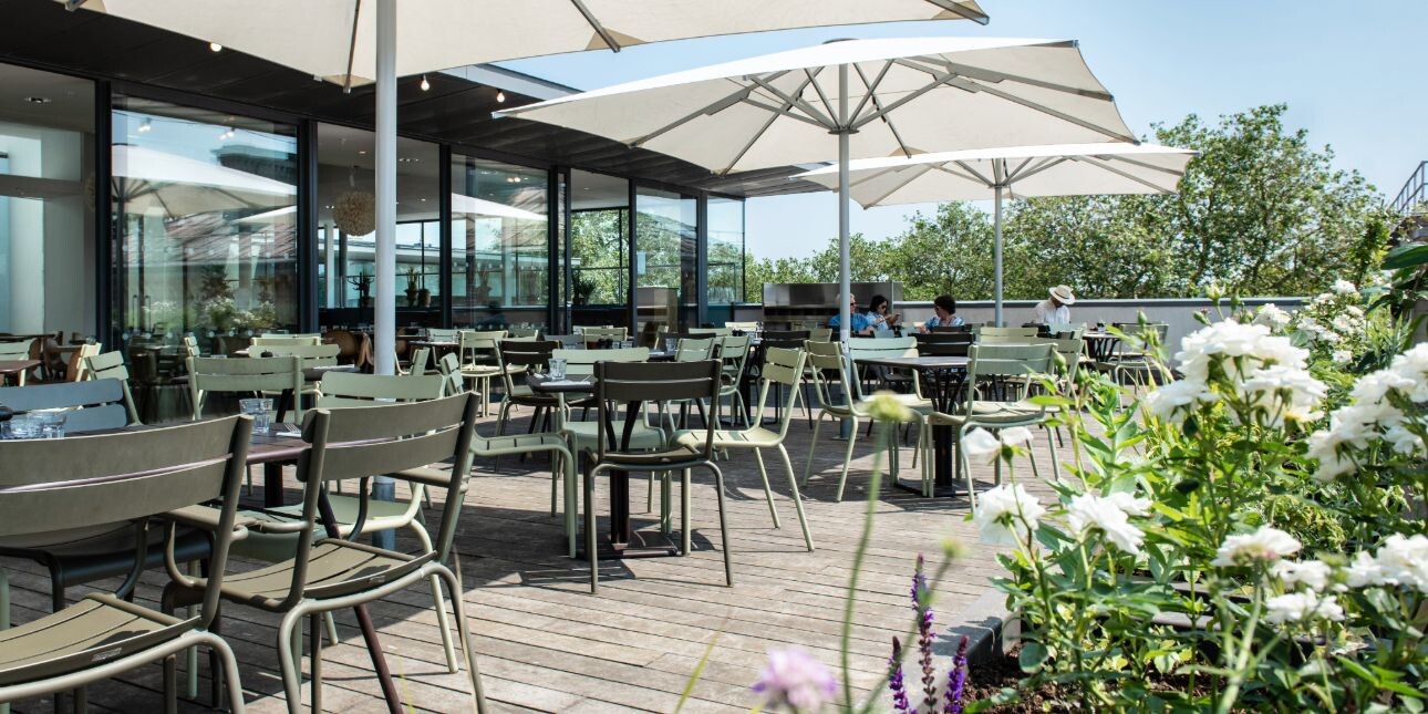 A restaurant roof terrace on a sunny day with green chairs, large white parasols, flower beds and a blue sky.