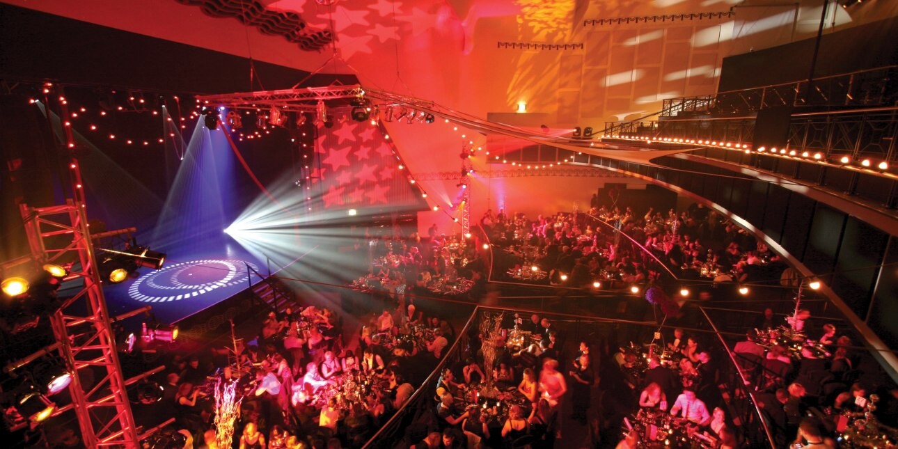 A colourful photo taken from a balcony overlooking the large auditorium and stages were audiences are sat around dozens of tables.