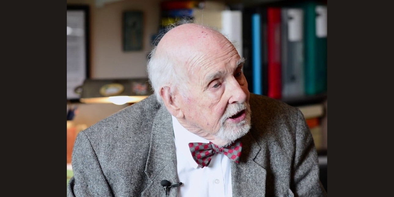 A colour portrait of Tim Travis-Healy looking off camera to his left. He is a white man with grey jacket and maroon bowtie.