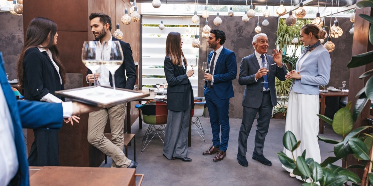 Five professionals of different ages holding wine glasses while networking in a modern event space