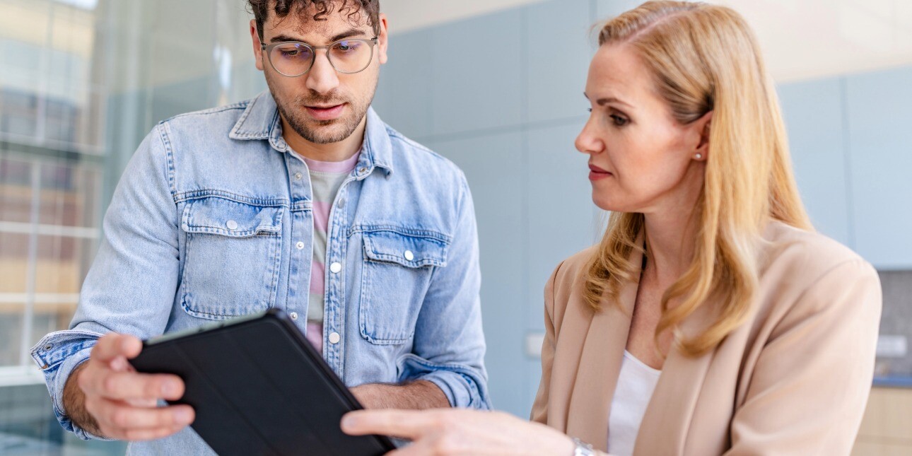 In a bright office, a young white man with dark curly hair and blue shirt holds a tablet while in conversation with a more senior female white colleague with blond hair and beige jacket