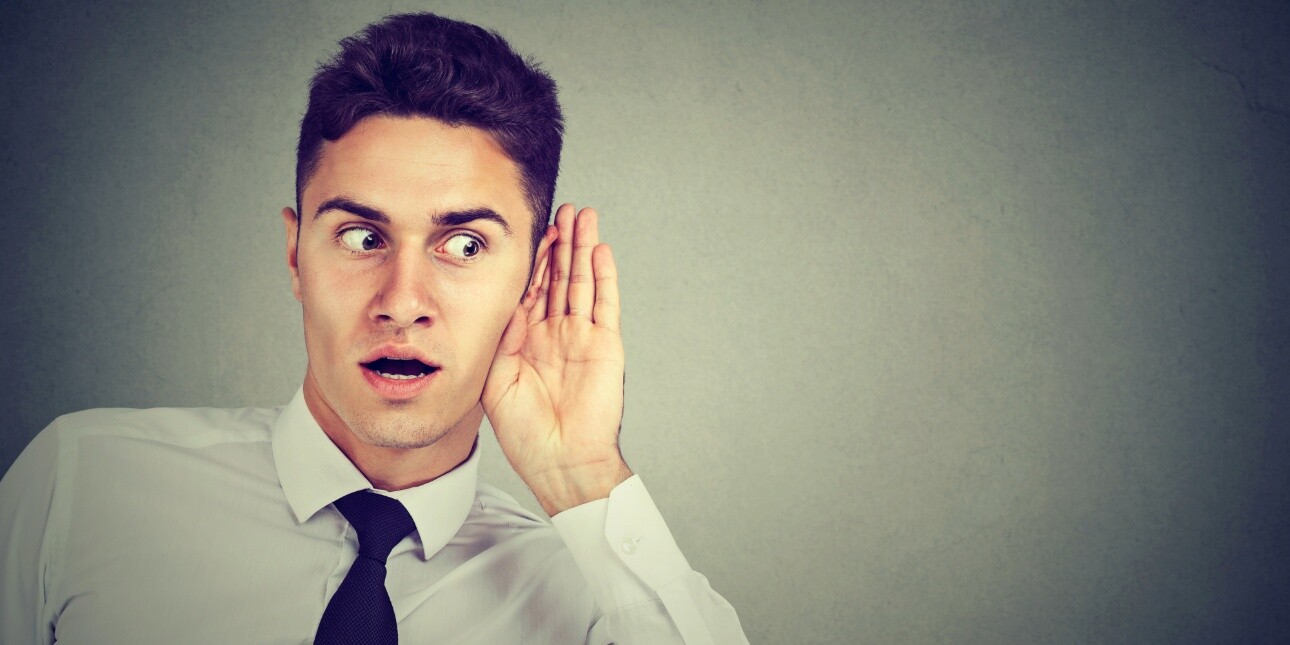 A young white man with dark hair, white shirt and black tie accentuates his features and looks to his left while cupping his left hand around his left ear.