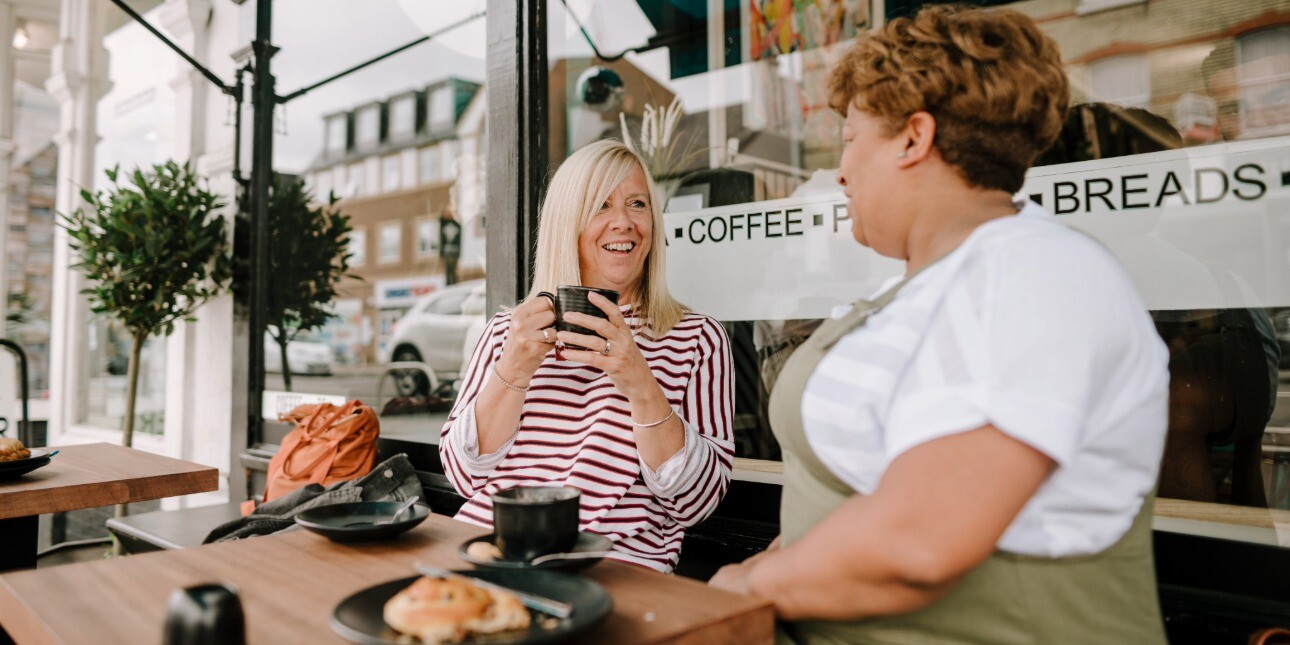 An older white woman with blonde hair and red striped top holds a coffee cup while sat outside a coffee shops chatting to a female member of staff in an apron.