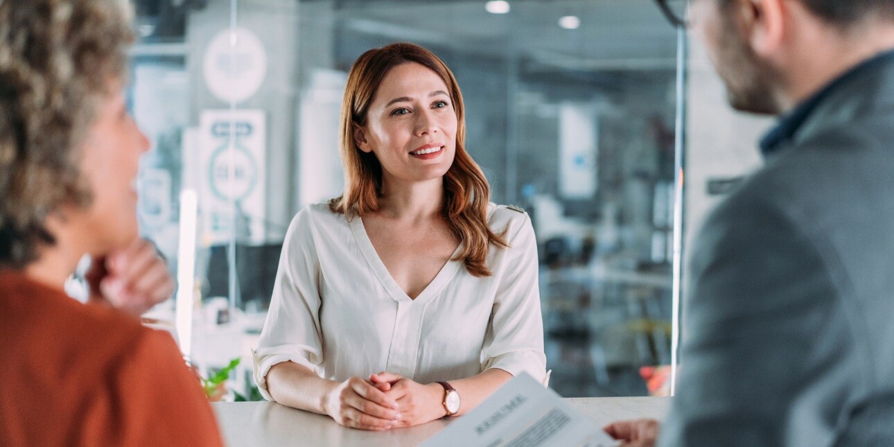 A white woman with brown hair and white top smiles during a job interview in a modern office. She is being interviewed by a woman in an orange top and a man in a grey suit. Both are partially visible in the shot.