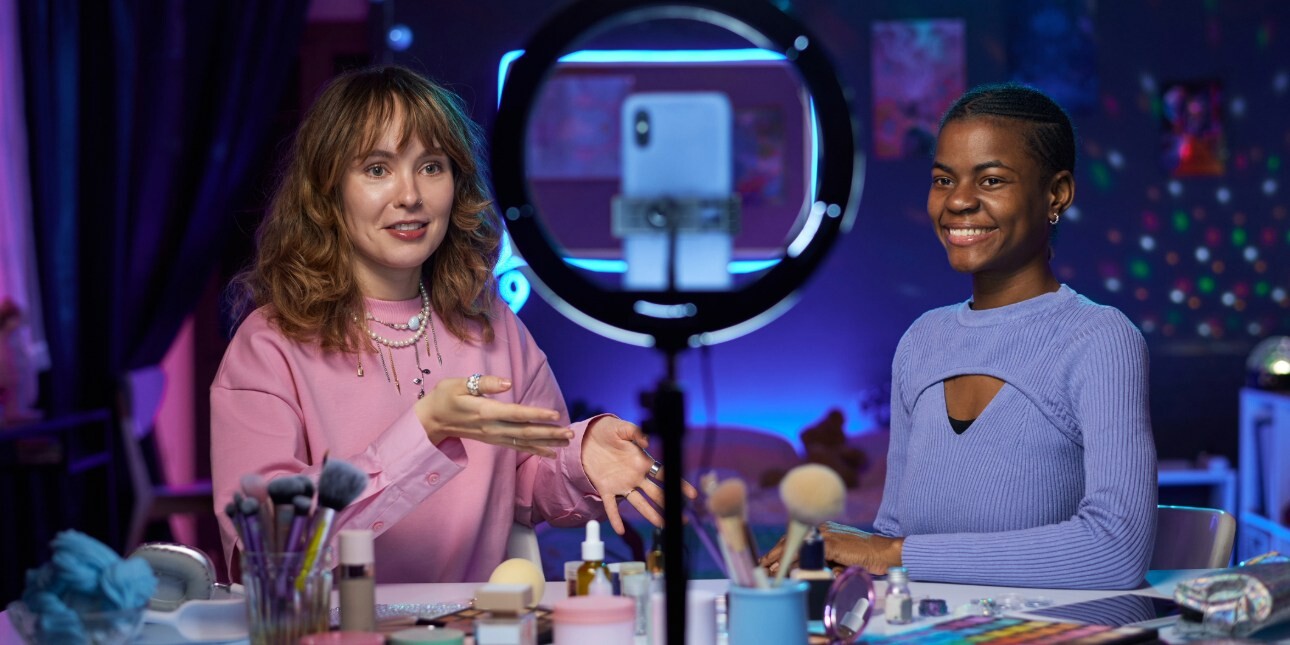 In a dark room with blue and pink neon hues a young white woman and a young Black woman sit facing a phone and ring flash demonstrating make up and beauty items which are on table in front of them.