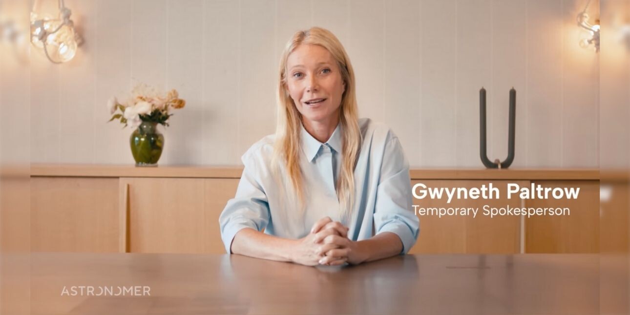 A screenshot featuring Gwyneth Paltrow - a white woman with long blond hair and pale blue shirt - sat at a wood table. Behind her are flowers and a candle holder on a wooden sideboard.