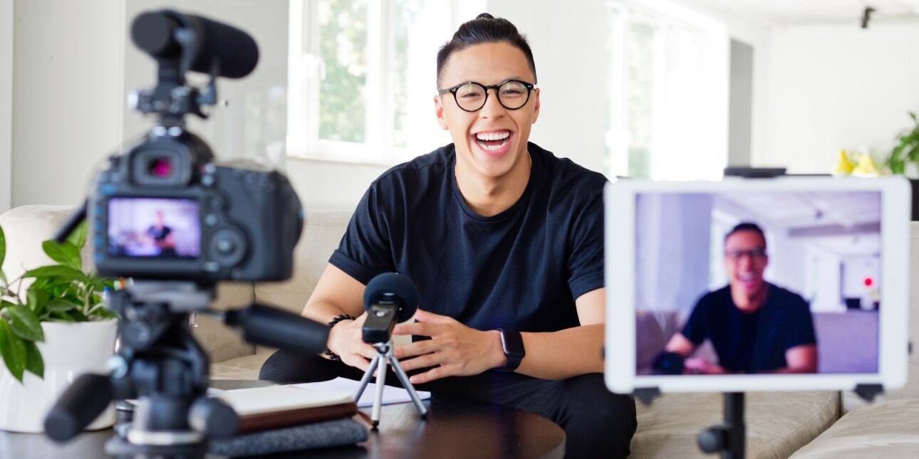 A smiling Asian man with short dark hair and dark shirt sits on a sofa in a modern home looking towards a camera and phone which are held on tripods..