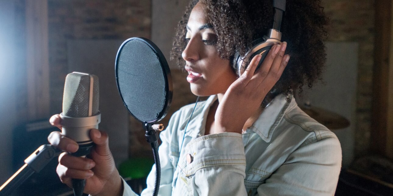 A young Black woman with shoulder length hair, headphones and blue denim jacket sings through a pop filter into a microphone that she is holding