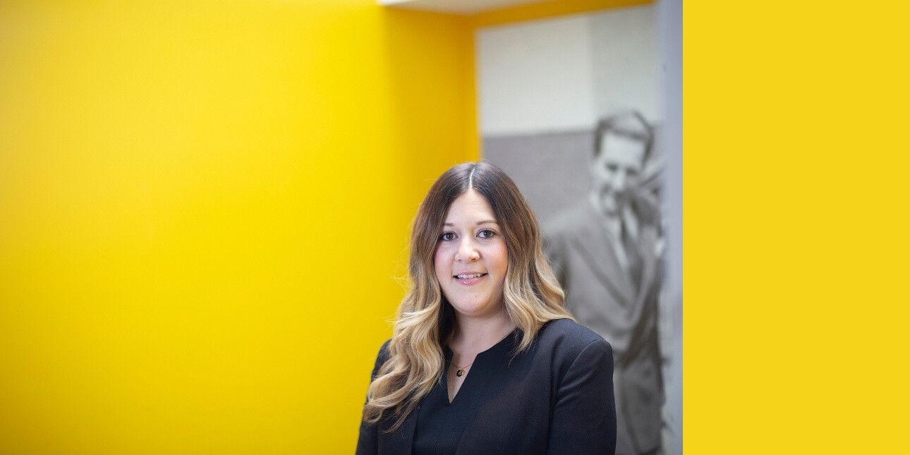 On a yellow background, a colour portrait of Laurien Hubbard. Laurien is a white woman with long, light brown hair who is wearing a black jacket. Behind her is a tall black and white photograph of a man.