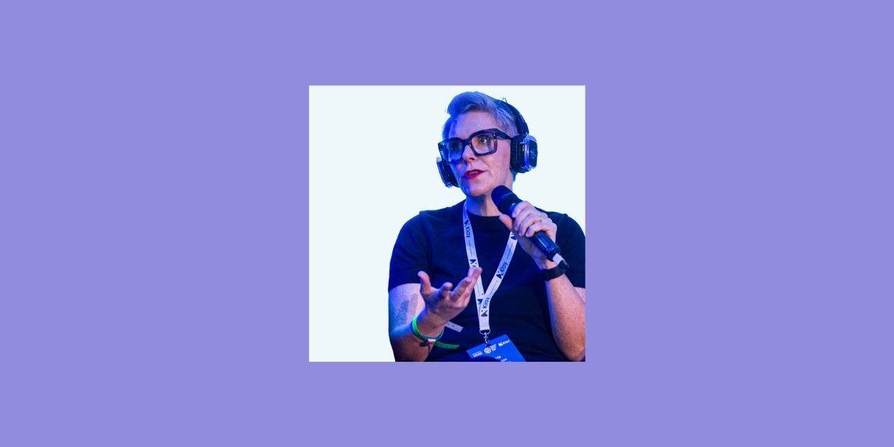 On a purple background, a colour photo of Orla Pearson on stage holding a microphone. Orla is a white woman with short blond hair.