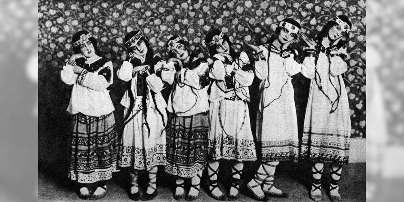 A black and white photograph of six dancers in embroidered tunics, long dresses and boots laying their heads on their hands