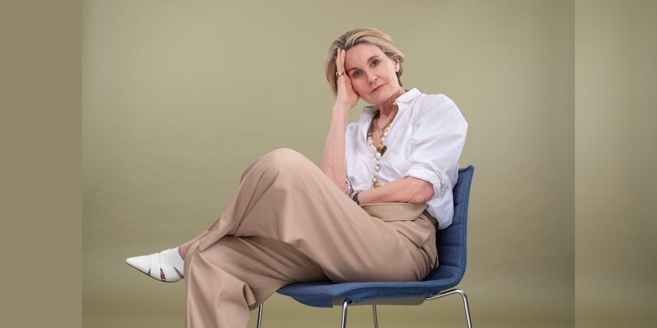 A colour photograph of Mary Portas sat in a chair against a beige background. Mary is a white woman with short blond hair wearing a white shirt and beige trousers.