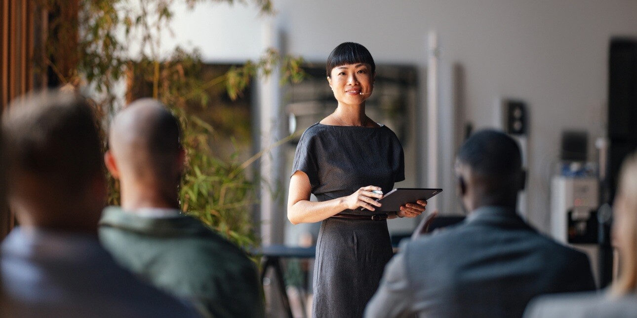 An Asian woman with short dark hair and grey dress looks at the camera while addressing an audience