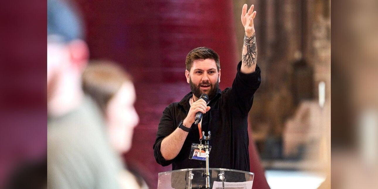A colour portrait of Daniel Coole in a conference setting holding up his left hand and holding a microphone in her right hand. Daniel is a white man with short dark hair and a tattoo on his arm. Two unidentified people are blurred in the foreground.