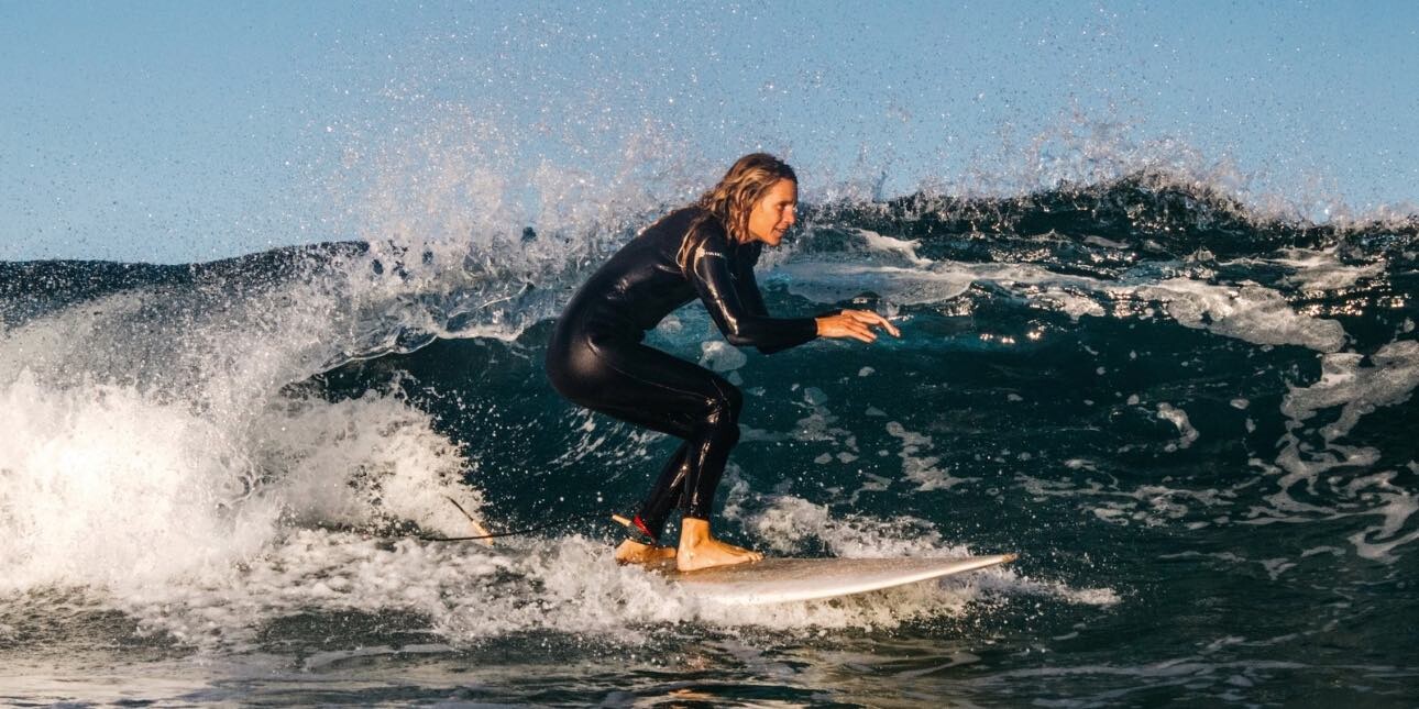A white female surfer wearing a black all body wetsuit surfing along calm waves with a blue sky behind her