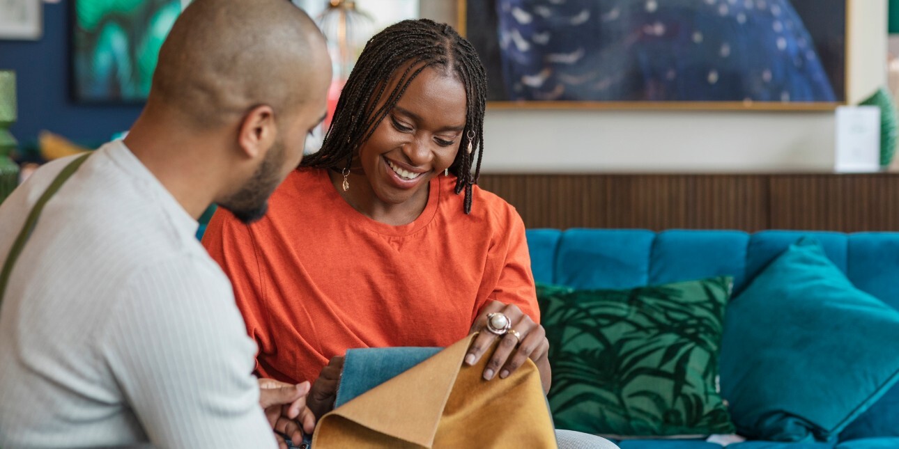A male and female black couple sat in a furniture shop choosing sofa fabrics. They are both black; the man wears a grey top and the woman wears an orange top.