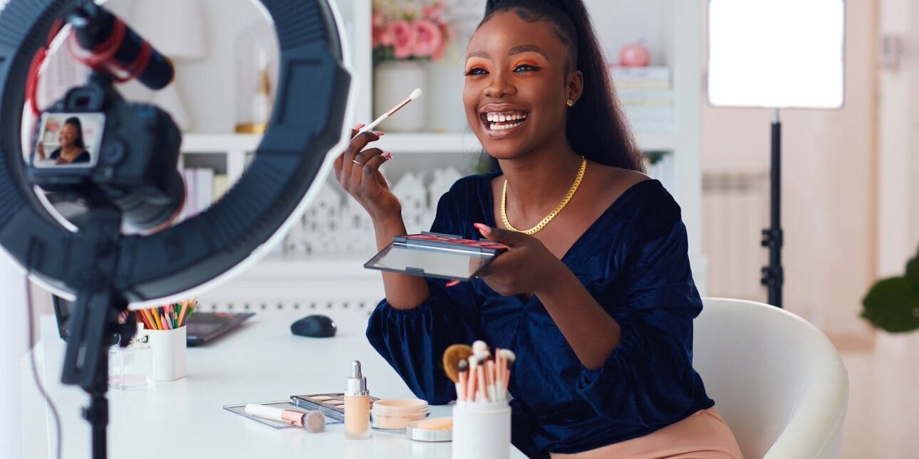 A smiling beauty-influencer holding up a make-up brush at the camera which is mounted on a ring-flash. The influencer is a black woman wearing a blue top, sat at a table on which there is placed other make-up accessories