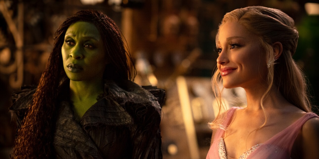 A movie still showing Cynthia Erivo as the green witch Elphaba with long dark hair and black outfit stood next to Ariana Grande as Glinda, the white witch with long blond hair and pink corset.
