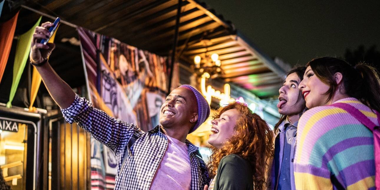 Friends in bright clothes taking selfies using a mobile phone outdoors at festival at night