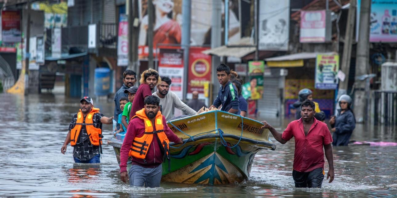 A group of rescuers wade through flooded streets as they pull a boat full of people they've rescued. There are buildings in the background.