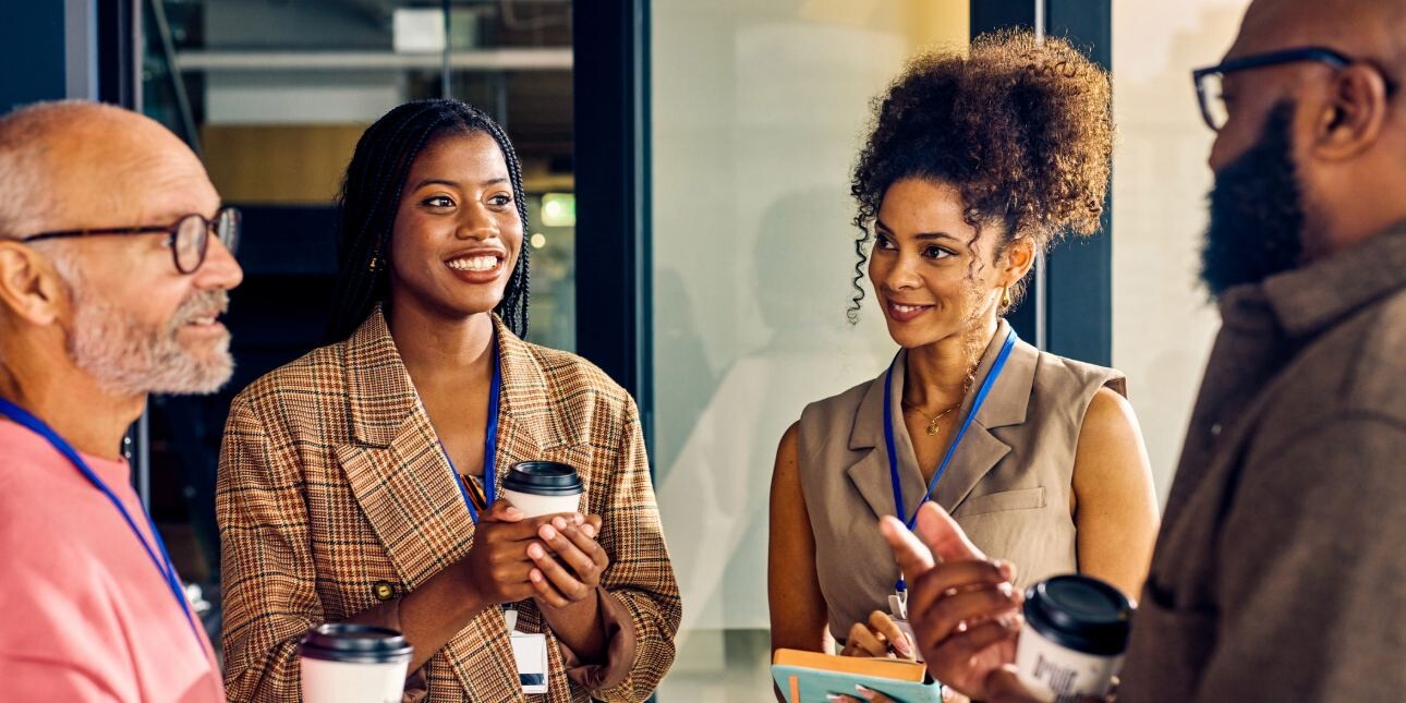 A group of four smiling colleagues of different ages stood holding coffee cups: a white man with grey beard, a Black woman with check jacket, a Black woman wearing an sleeveless top, and a Black man wearing a brown shirt.