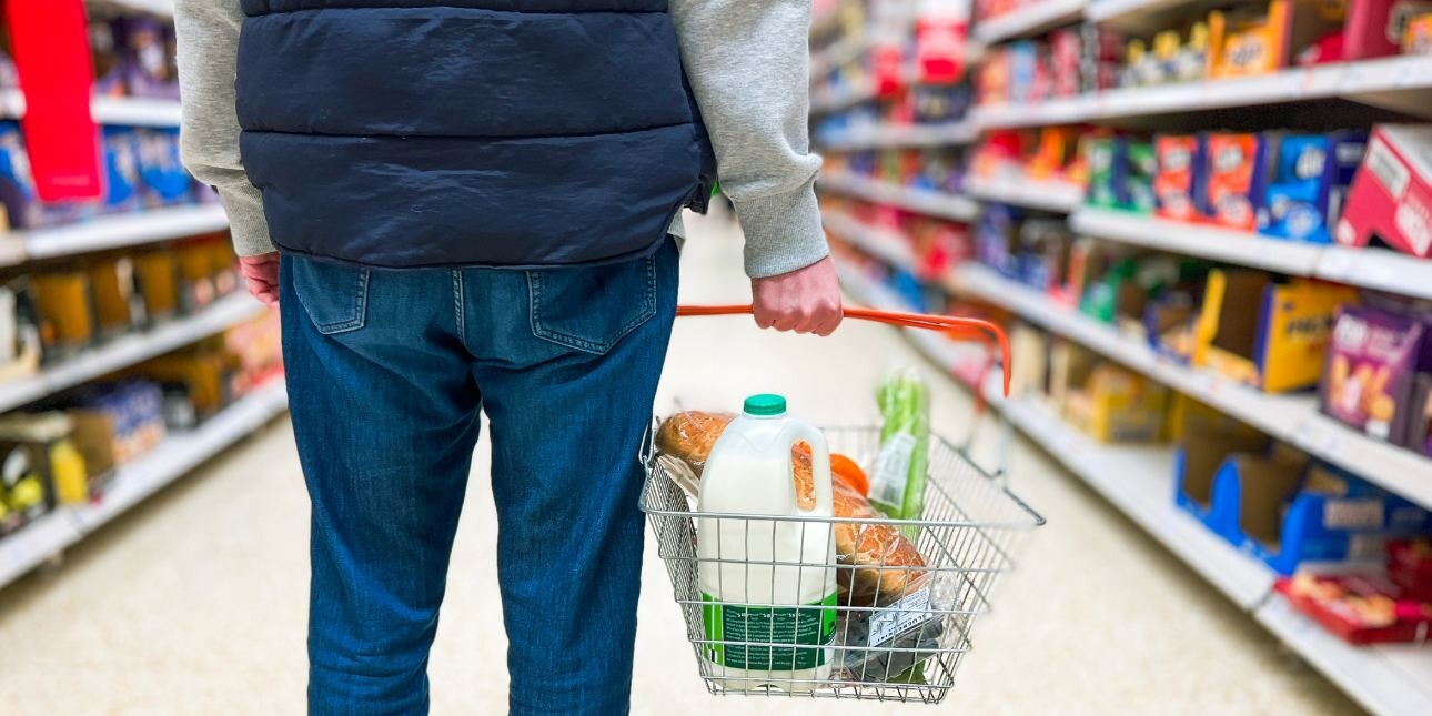 The back of a man in a supermarket aisle wearing a blue gilet and blue jeans holding a shopping basket full of bread, milk and groceries.