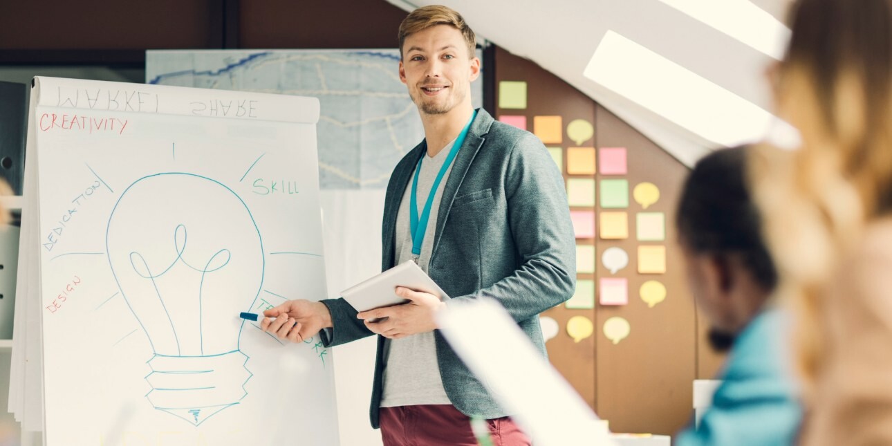 A smiling white man dressed in business-casual clothing stands in front of flip chart, holding a digital tablet, while addressing colleagues sat around a table.