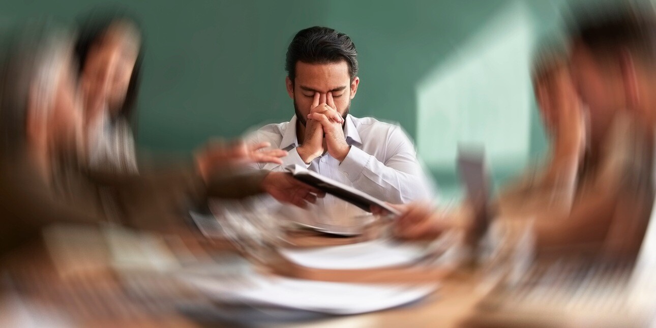 A concept of stress showing a business man holding his head in his hands while a motion blur shows colleagues around table handing him paperwork