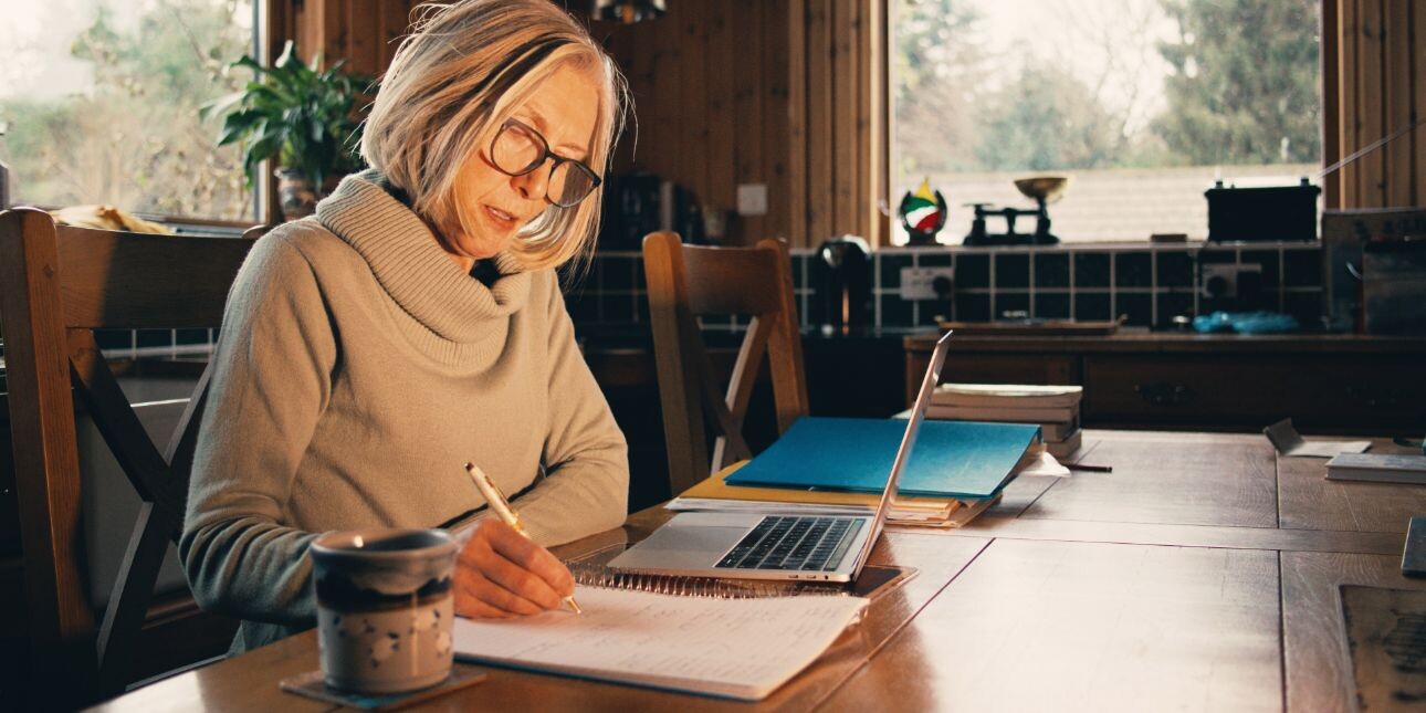 A middle aged white woman with a grey bob and beige jumper sat at a kitchen table writing in a notepad while surrounded by a laptop and paperwork.