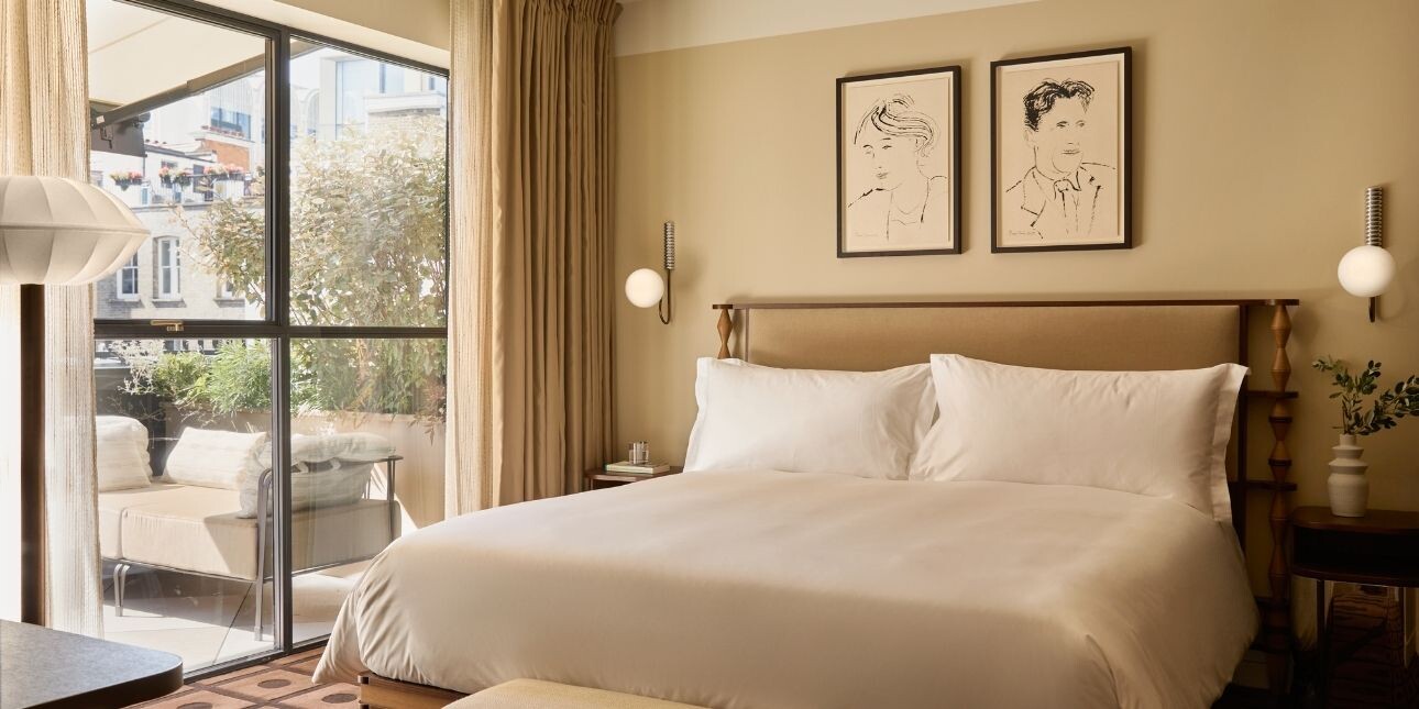 An exquisite hotel room furnished in cream shades with a large bed, pictures and doors to a terrace.