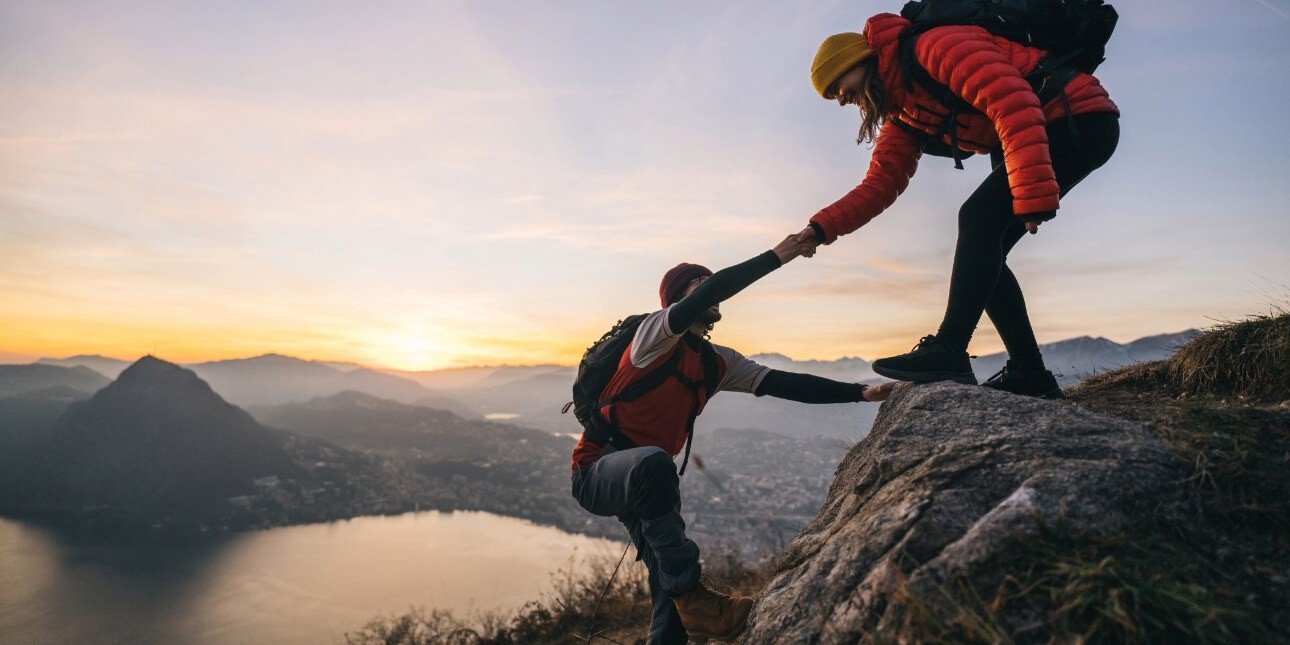 A female hiker offers a male hiker a hand up rocks on a mountain. A lake is visible in the distance below.