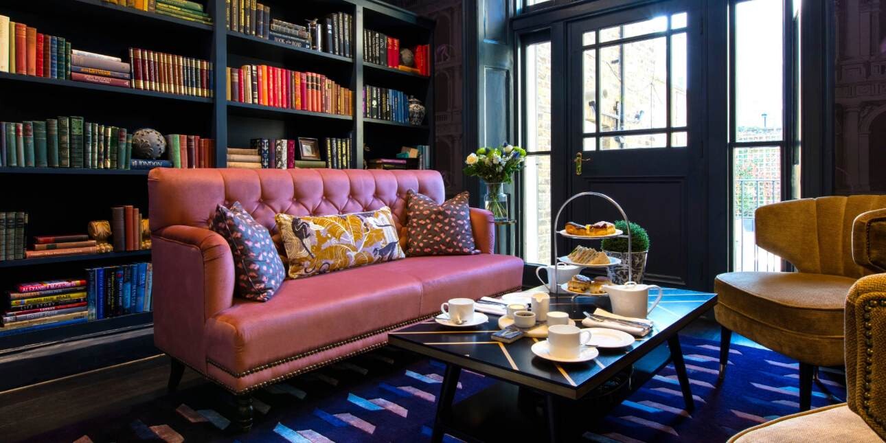 A dark blue room with a floor to ceiling bookcase, pink sofa and tan armchairs set around a coffee table with a traditional cream tea.