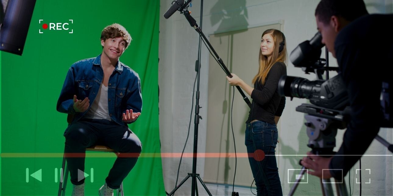 A picture of a young white man sat on a stool in a studio against a green screen. A white woman stood next to him holds a microphone boom while another man crouches behind a camera. The shot is overlaid with video control buttons.